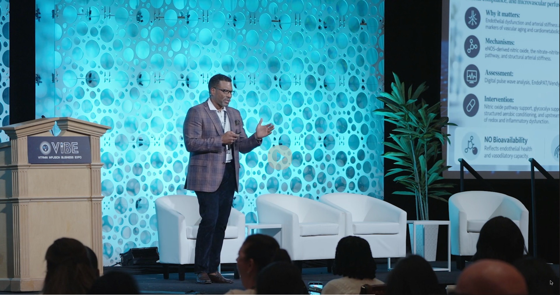 Dr. Christopher Davis presenting on stage at Vibe Expo 2026 in Las Vegas, gesturing toward the audience with his slide on endothelial health on screen beside him and the VIBE podium at his side