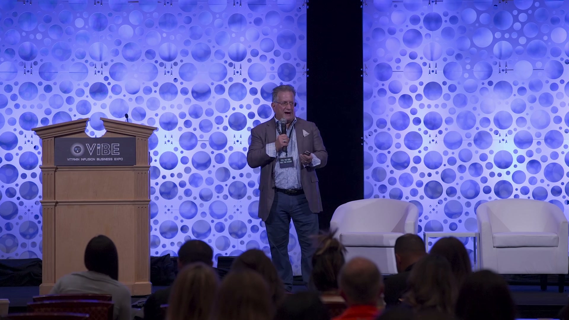 Dr. Mitch Ghen speaking on stage at Vibe Expo 2026 in Las Vegas, microphone in hand, with the conference audience seated in front of him